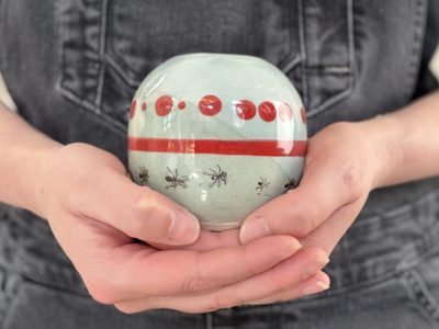 Hands holding a round vase with an ant illustration and red dots