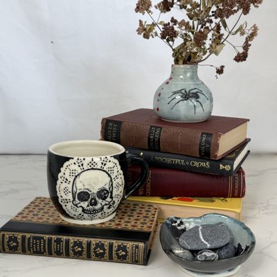Skull mug with lace border, books, and a bud vase with dried flowers